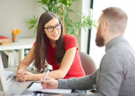 A man in a grey shirt is making notes while talking to a woman in red, both sat at a desk together.