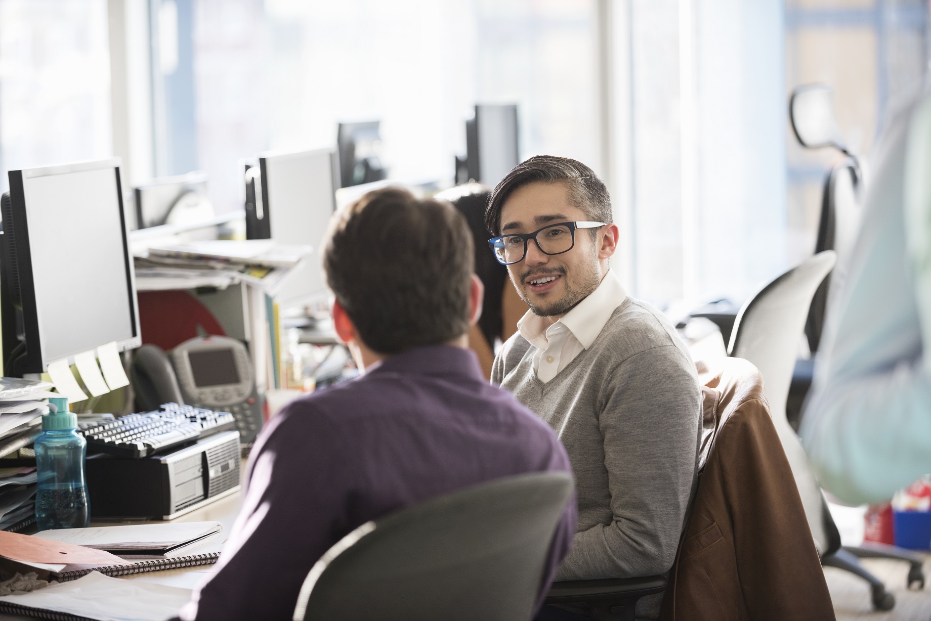 Two men in shirts sitting at a desk in front of computers talking to each other.