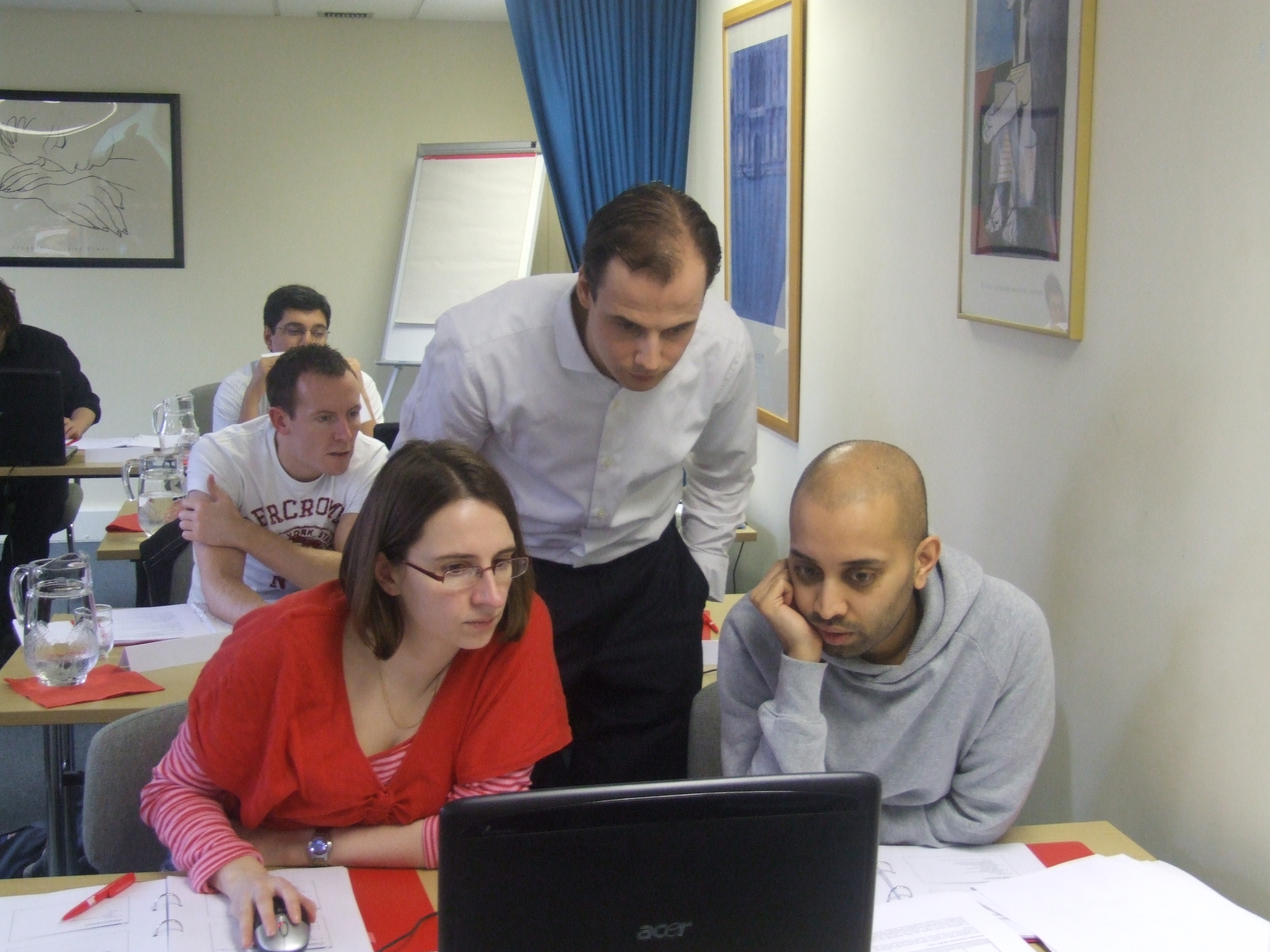Three people looking at a laptop in the foreground, more people are also looking at laptops on tables behind them.