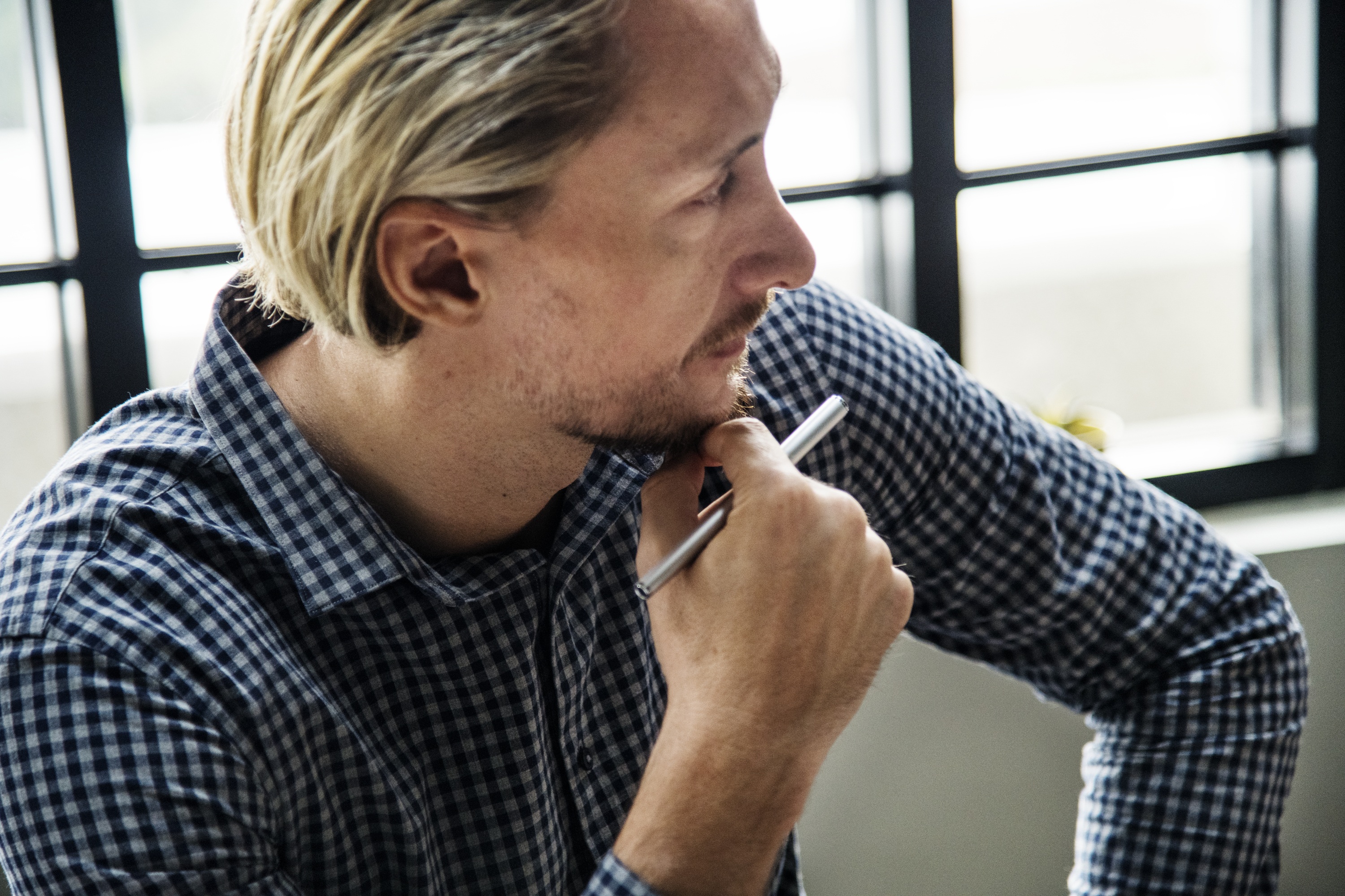 A man in a checked shirt holding his pen and looking thoughtfully off camera.