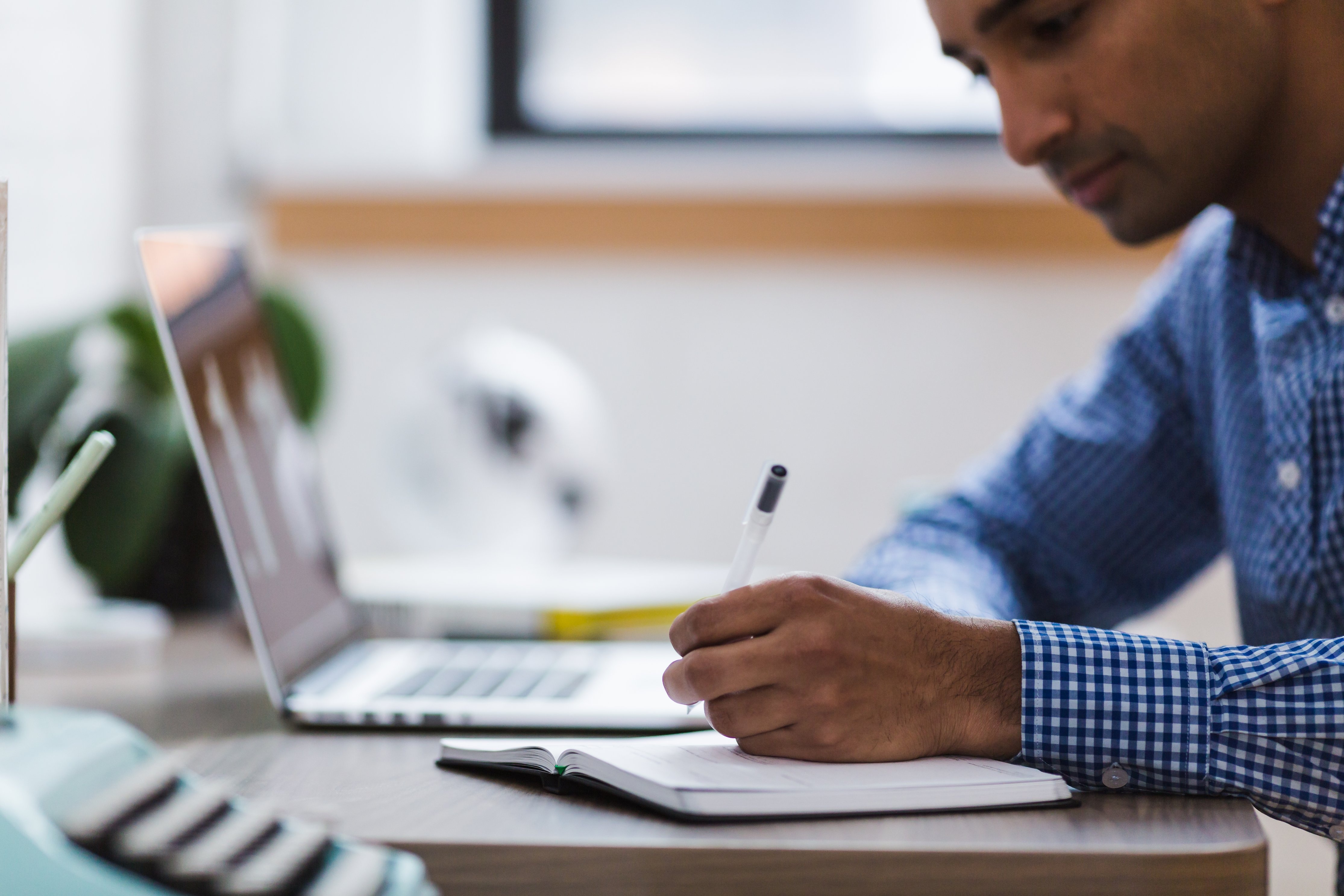 A man in a checked shirt writing in a notebook, with a laptop open on the desk in the background.