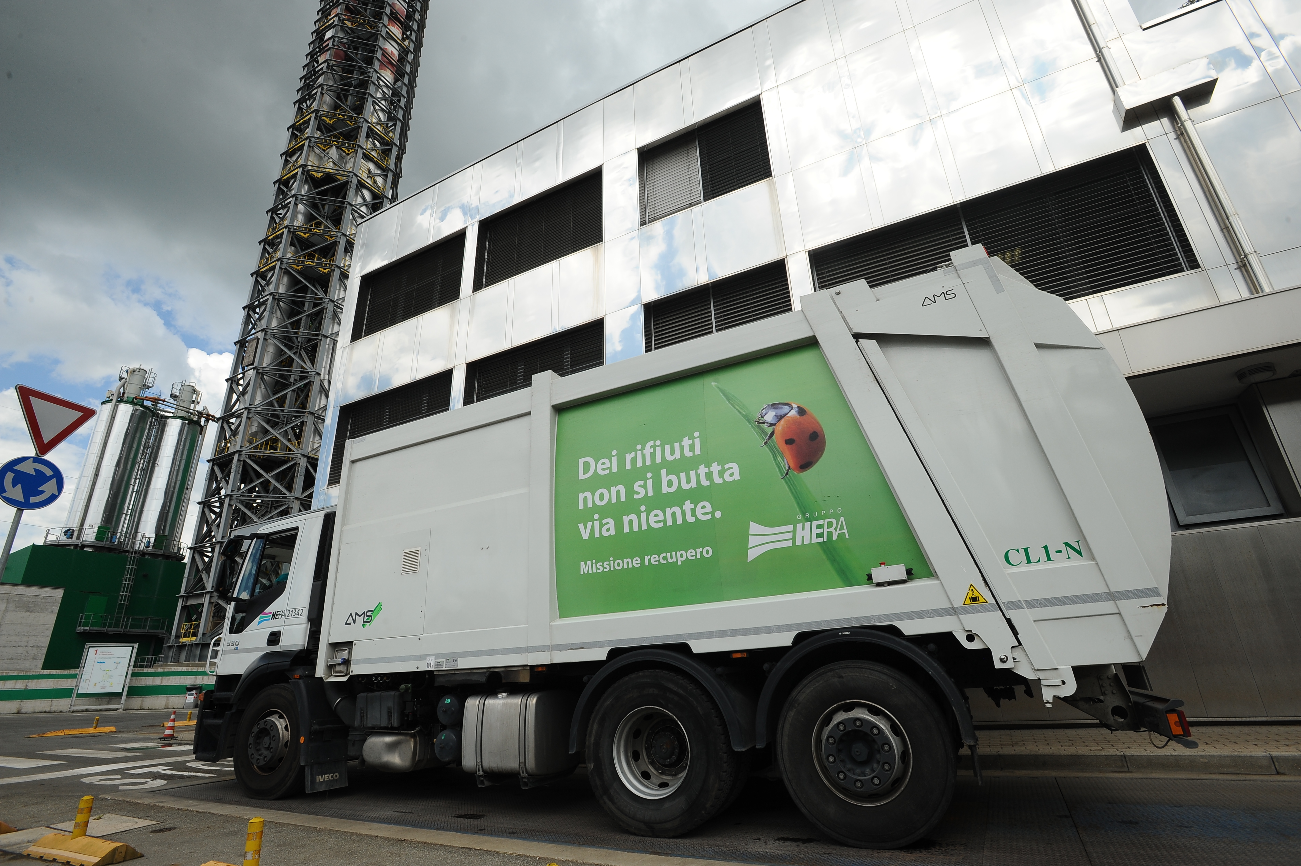 White Italian bin lorry stopped at a roundabout outside a large, factory building.