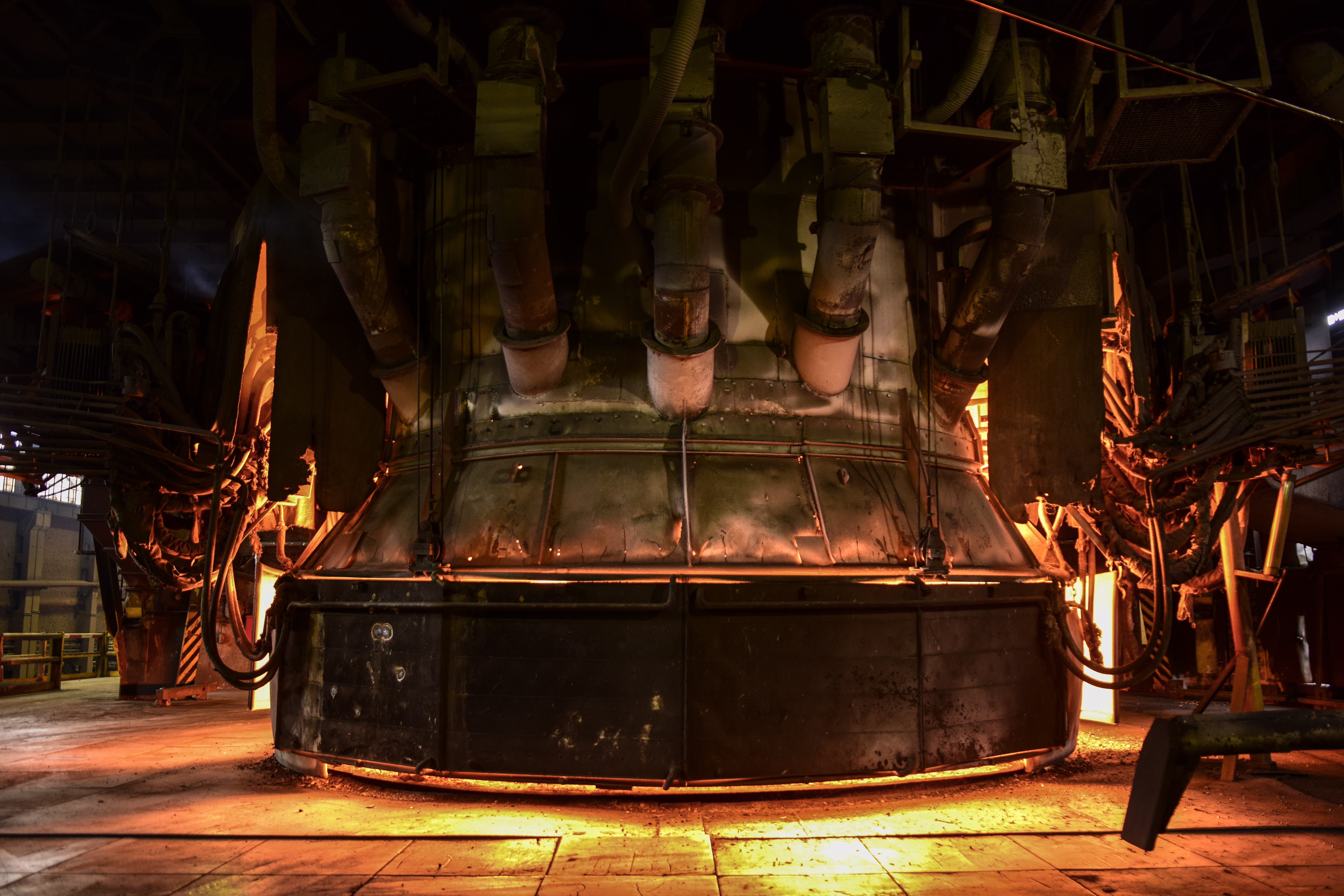 Interior of a factory featuring a large metallurgy machine, with an orange glow coming from around and inside it.