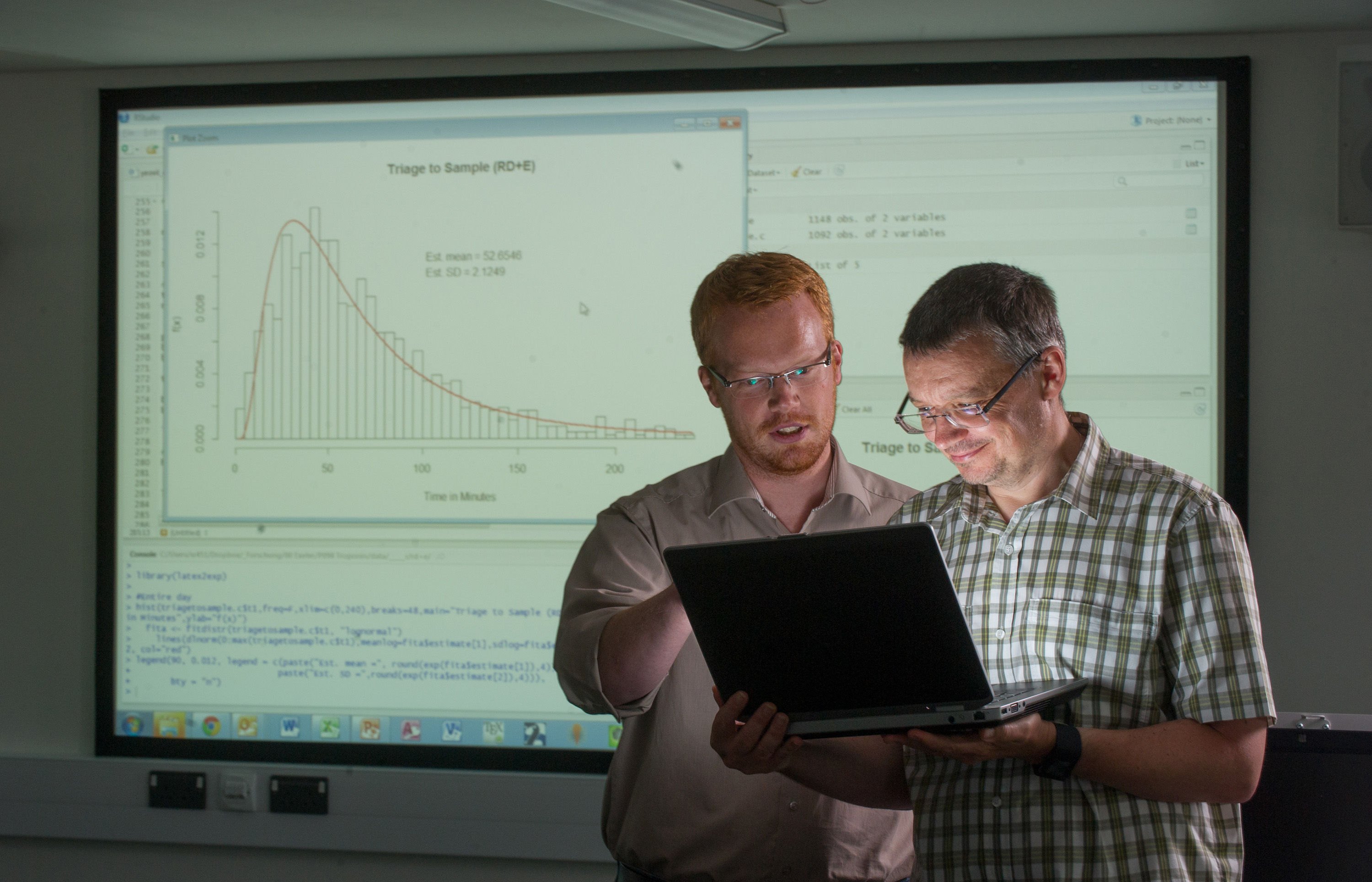 Two men in shirts looking at a laptop in front of a projector with a graph on the screen.
