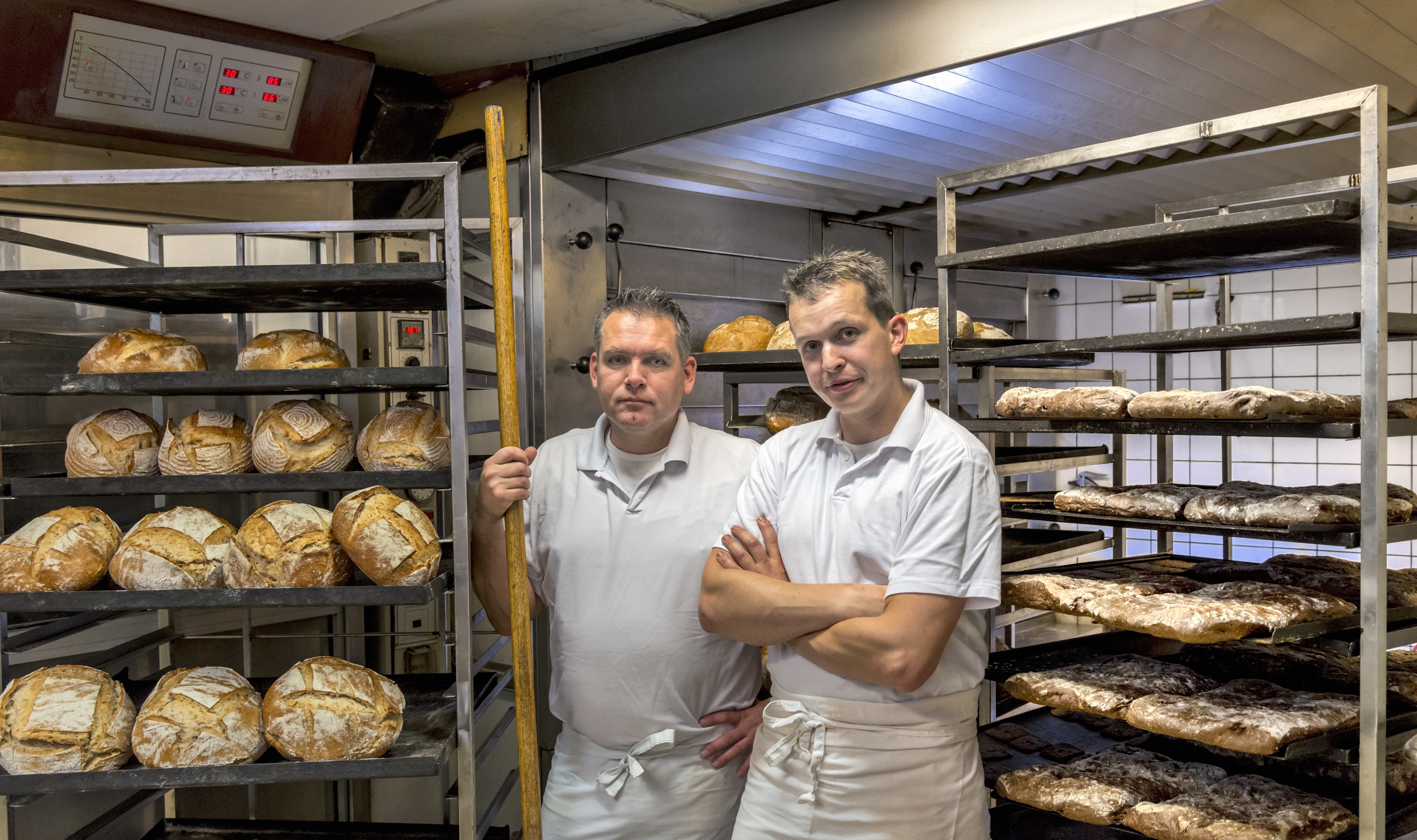 Two bakers in white polo shirts with aprons round their waists posing next to their latest batch of loaves on shelves.