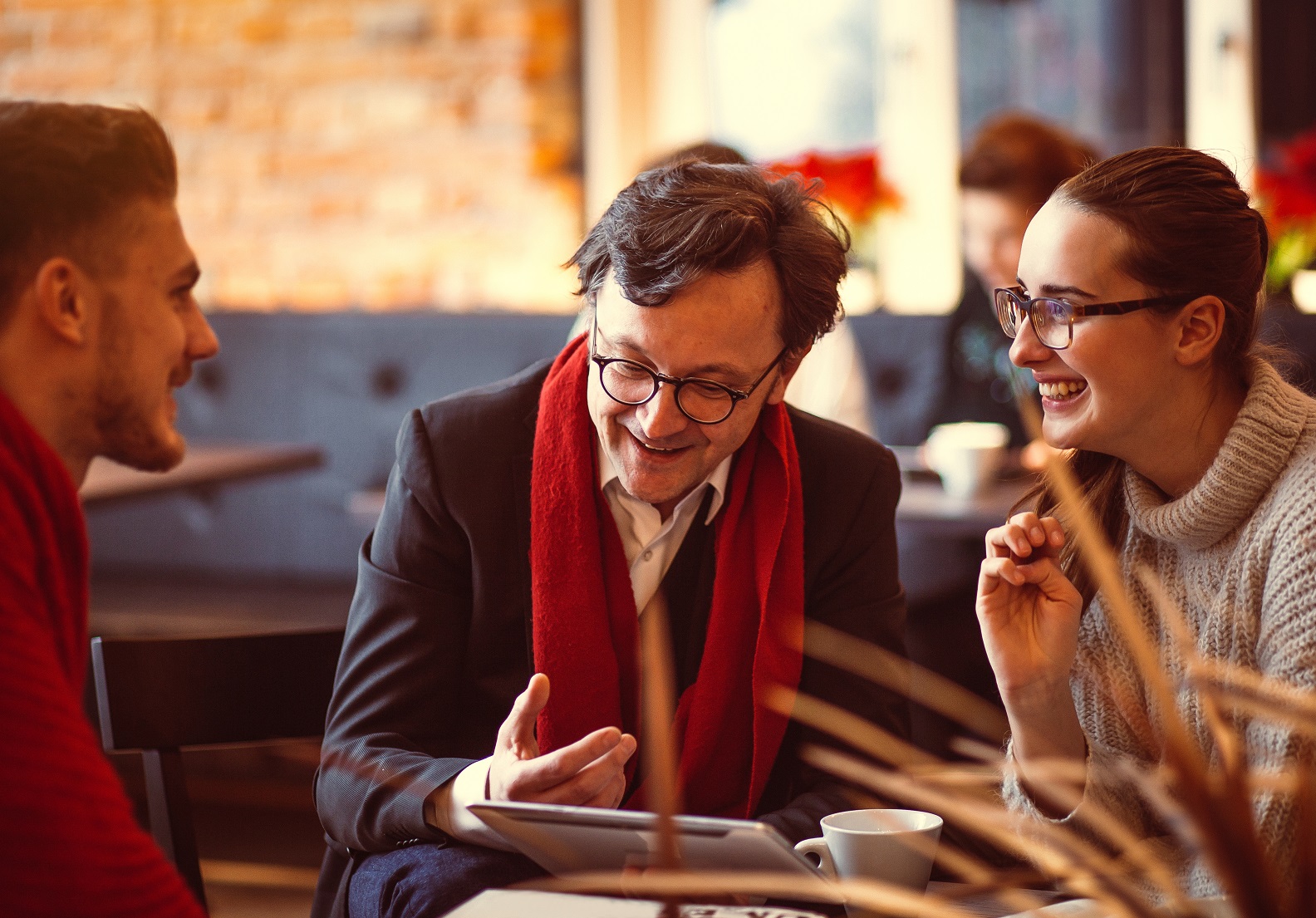Two men and a woman sat at a coffee table talking and laughing together while holding a tablet computer.