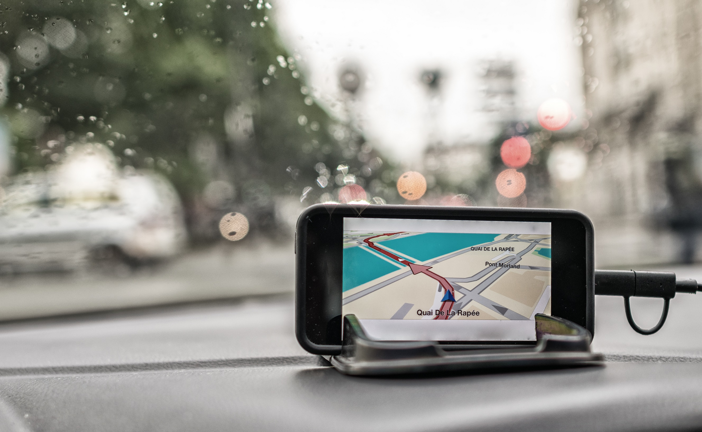 iPhone on a stand on the dash of a car, with a sat nav guidance app running on it directing the car, rain is on the window screen behind the phone.