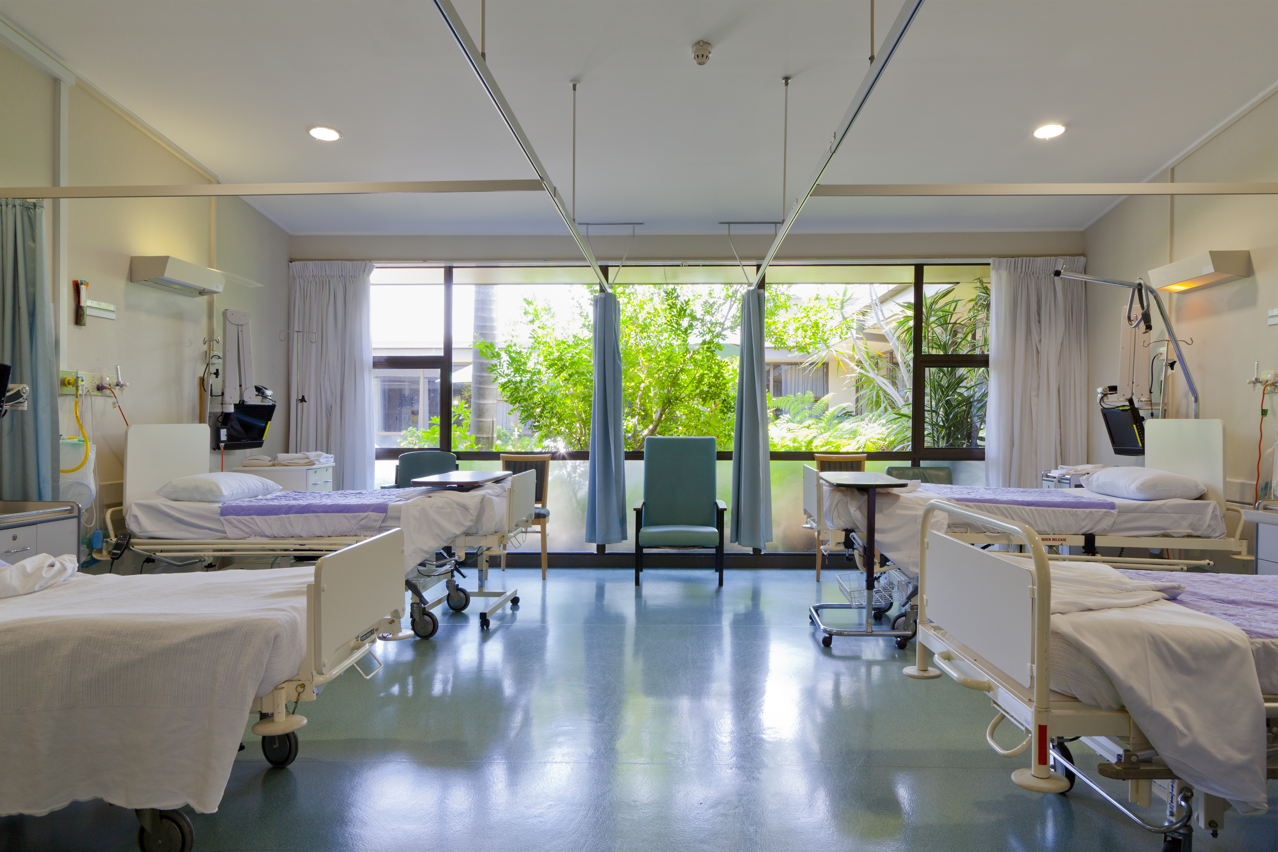 An empty hospital room with four beds and some chairs inside, with large windows looking out onto some greenery outside.