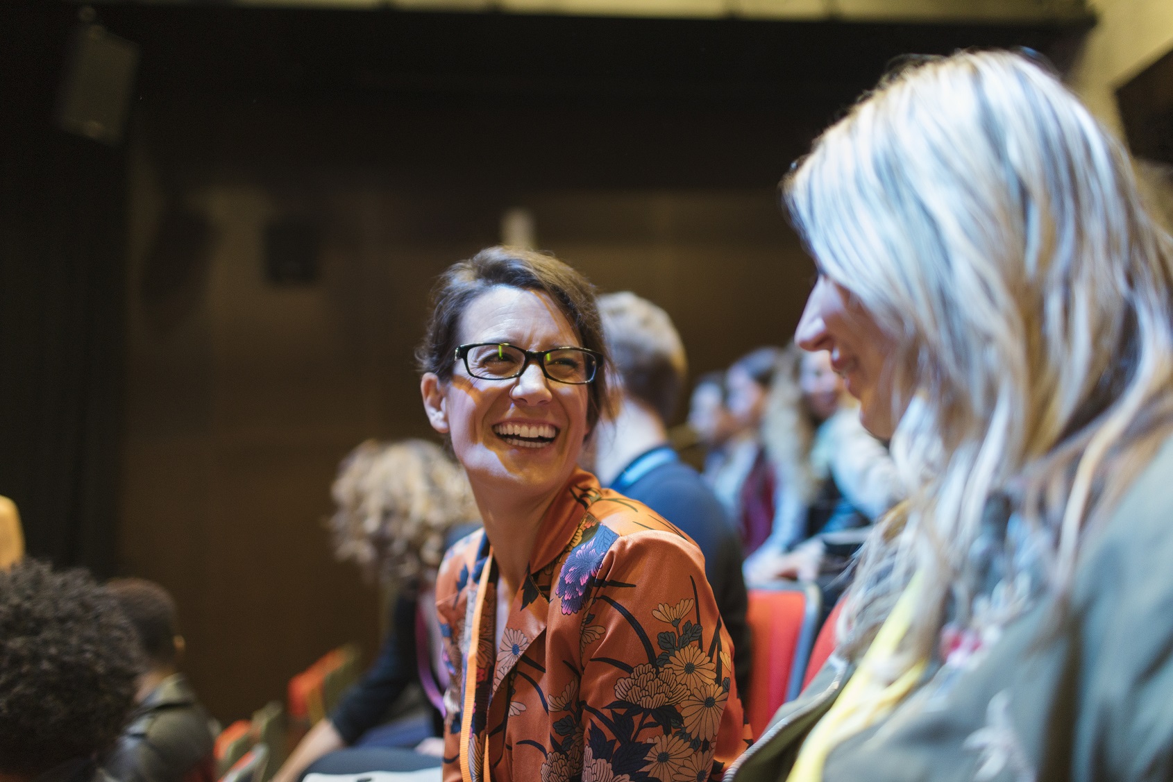 Two women in lecture hall audience share a laugh